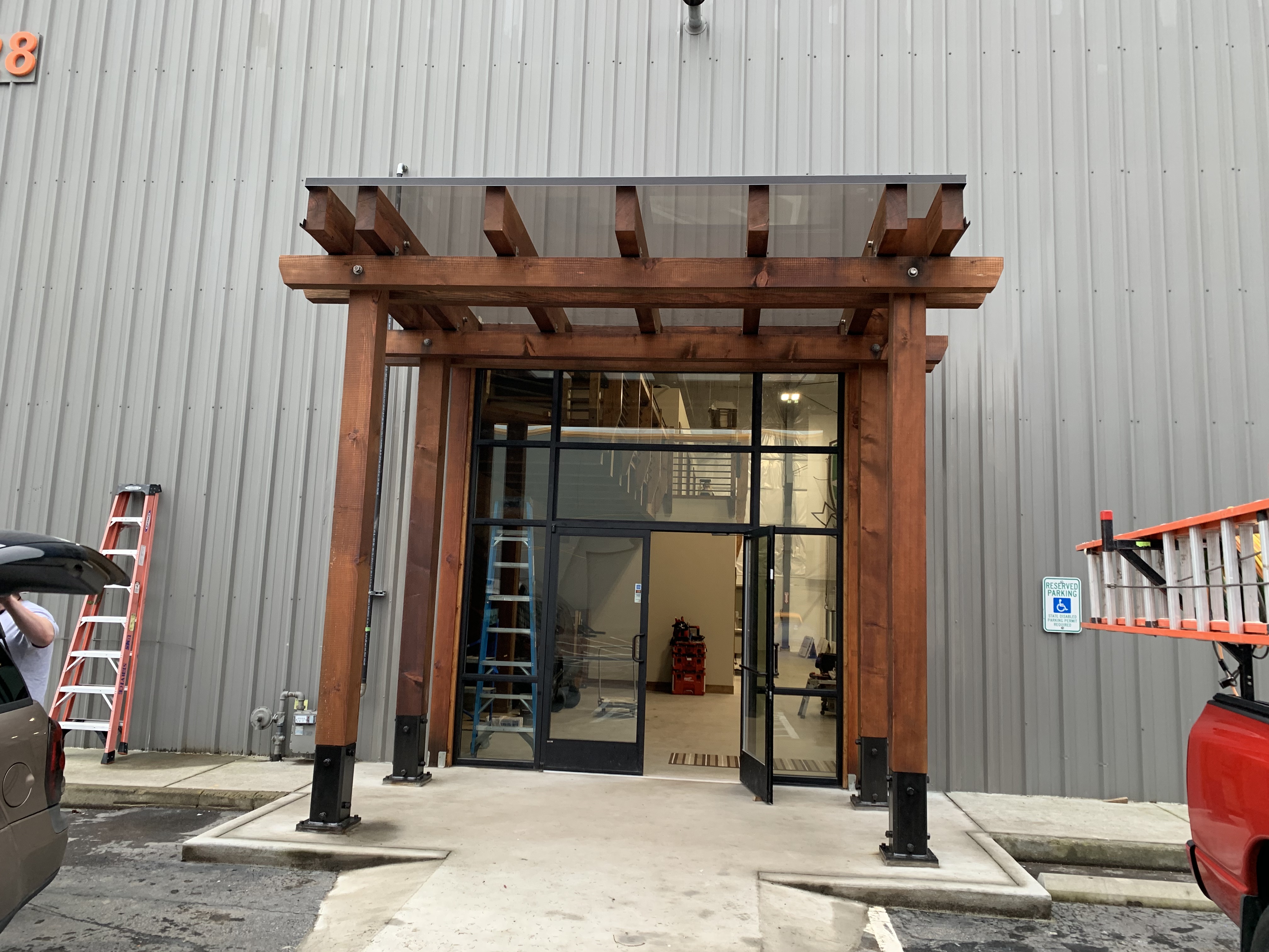 Stained timber entry pergola with a glass canopy over a storefront door on a metal building