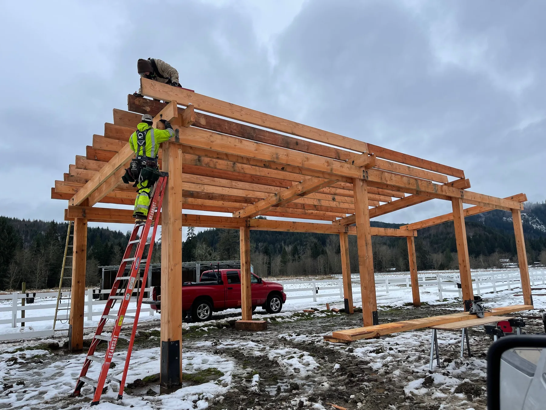 Douglas Fir timber frame pavilion being raised in a snowy pasture, crew setting the ridge beam from ladders