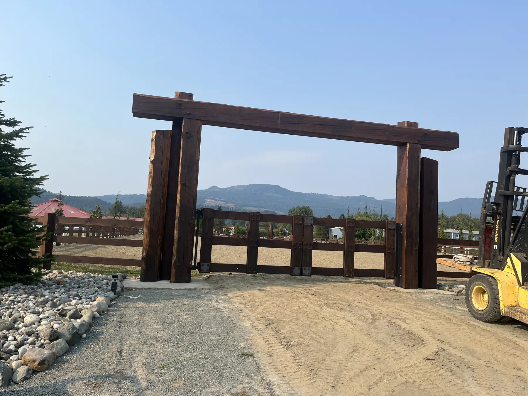 Heavy timber entry gate with dark stained Douglas Fir posts and a long header beam, mountain view and forklift behind